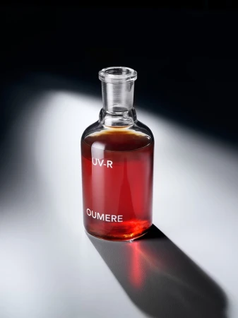 Laboratory flask with dark amber liquid on a white surface. Shadow cast behind the flask. White text 'UV-R' and 'OUMERE' on bottle.