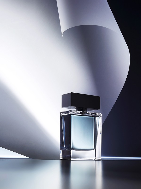Elegant product shot: a glass perfume bottle with a black cap stands against a backdrop of curved white paper in a photo studio setting.