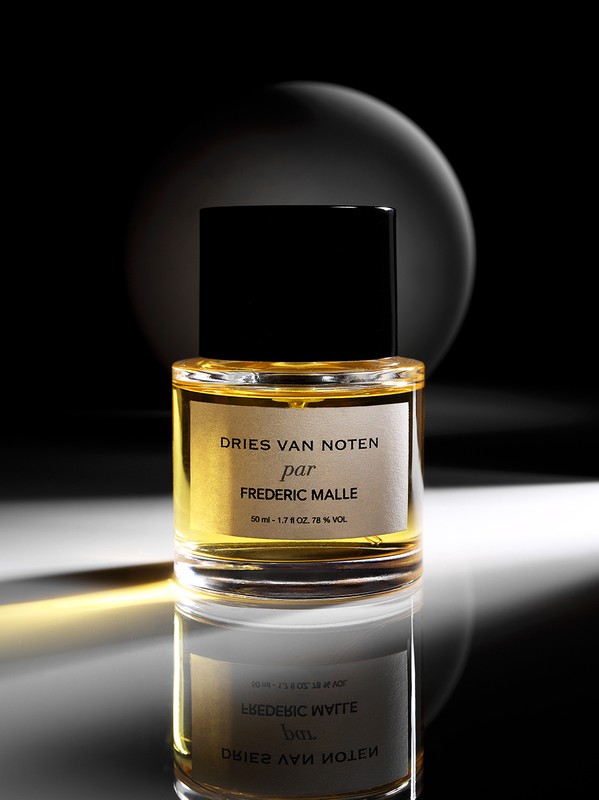 Close-up studio shot of a Dries Van Noten par Frederic Malle perfume bottle on a reflective surface against a dark background with light orb.