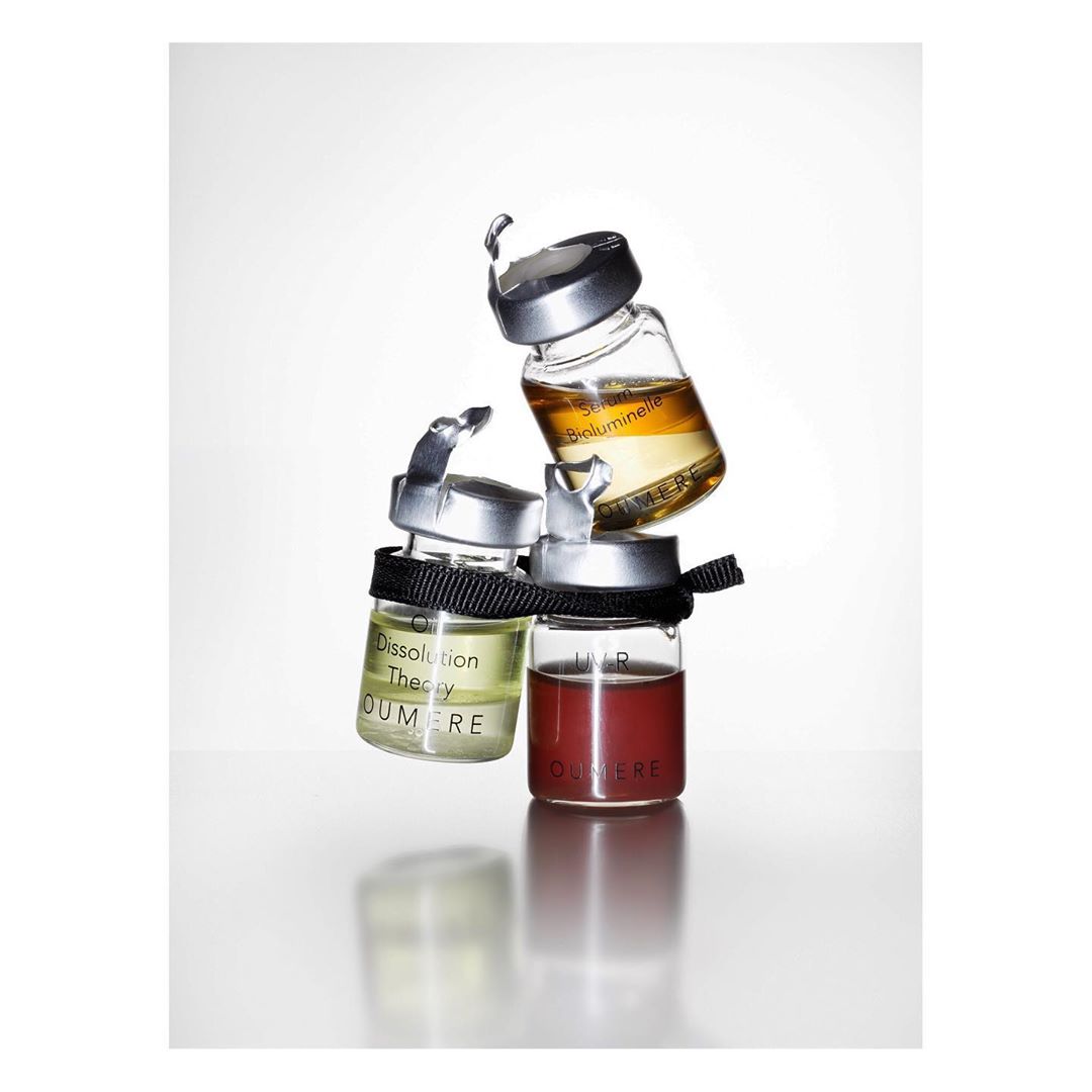 Three small clear bottles of cosmetic serums stacked on top of each other with black ribbon around the bottle necks on white.
