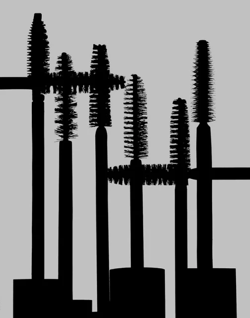 Multiple mascara brushes are arranged closely together on a gray background, creating a dense and textured composition in black and gray tones.