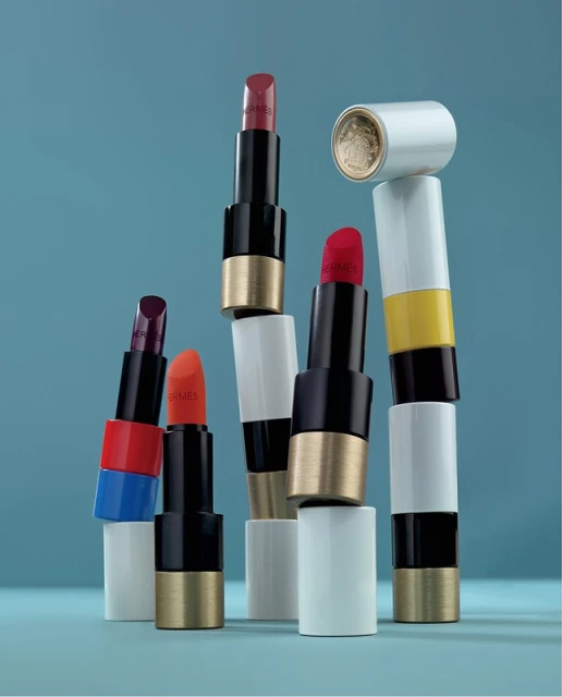 Arrangement of colourful Hermes lipstick tubes against a blue background, with varied shades and textures creating a beauty still life.