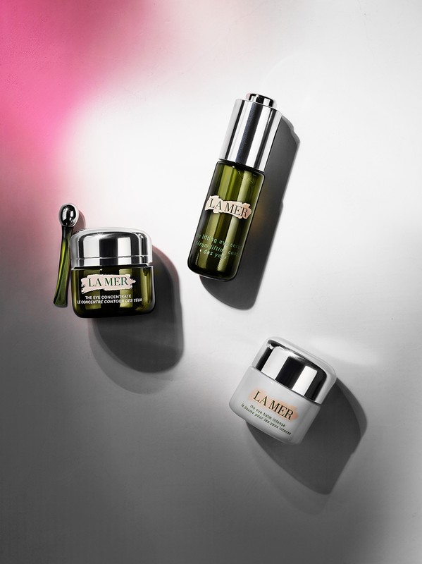 Still life shot of three La Mer skin care products: a serum, an eye balm, and a face cream, arranged against a split pink and grey background.