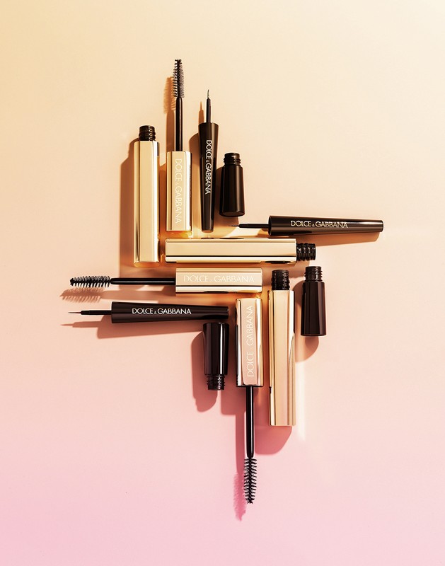 Several Dolce & Gabbana black and gold mascara tubes and applicators arranged on a gradient peach background, beauty and cosmetics still life.