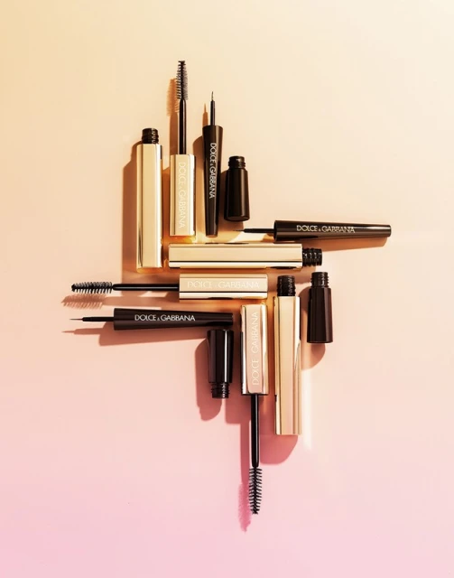 Several Dolce & Gabbana black and gold mascara tubes and applicators arranged on a gradient peach background, beauty and cosmetics still life.