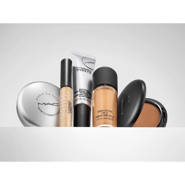 Studio shot of MAC makeup products including foundation, concealer, and powder compacts, arranged on a white surface against a grey background.