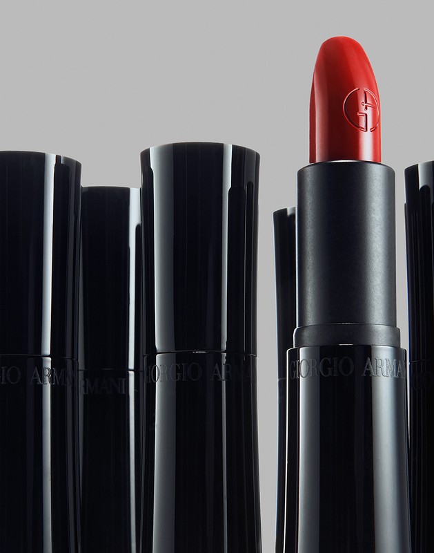 Close-up product photograph of a bright red lipstick tube standing out among several black capped lipsticks against grey