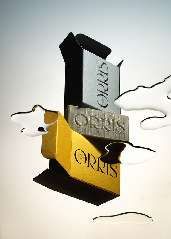 Three bars of Orris soap stacked in a tower, surrounded by pools of water on a gradient white to grey background in studio lighting.