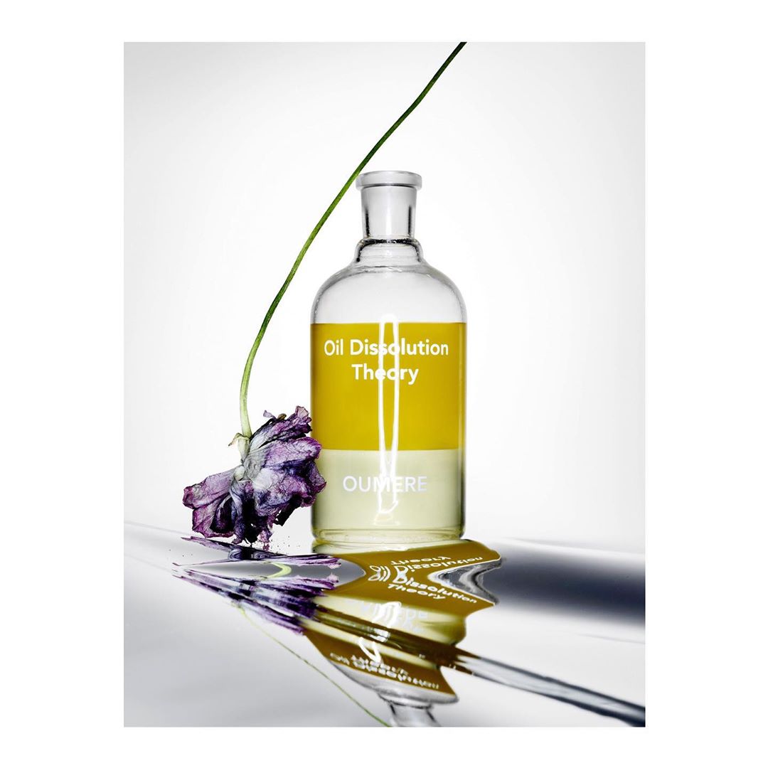 Still life product shot: A bottle of Oil Dissolution Theory with a purple flower, reflecting on a silver surface, against white.