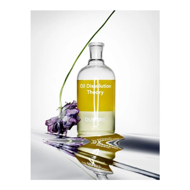 Still life product shot: A bottle of Oil Dissolution Theory with a purple flower, reflecting on a silver surface, against white.