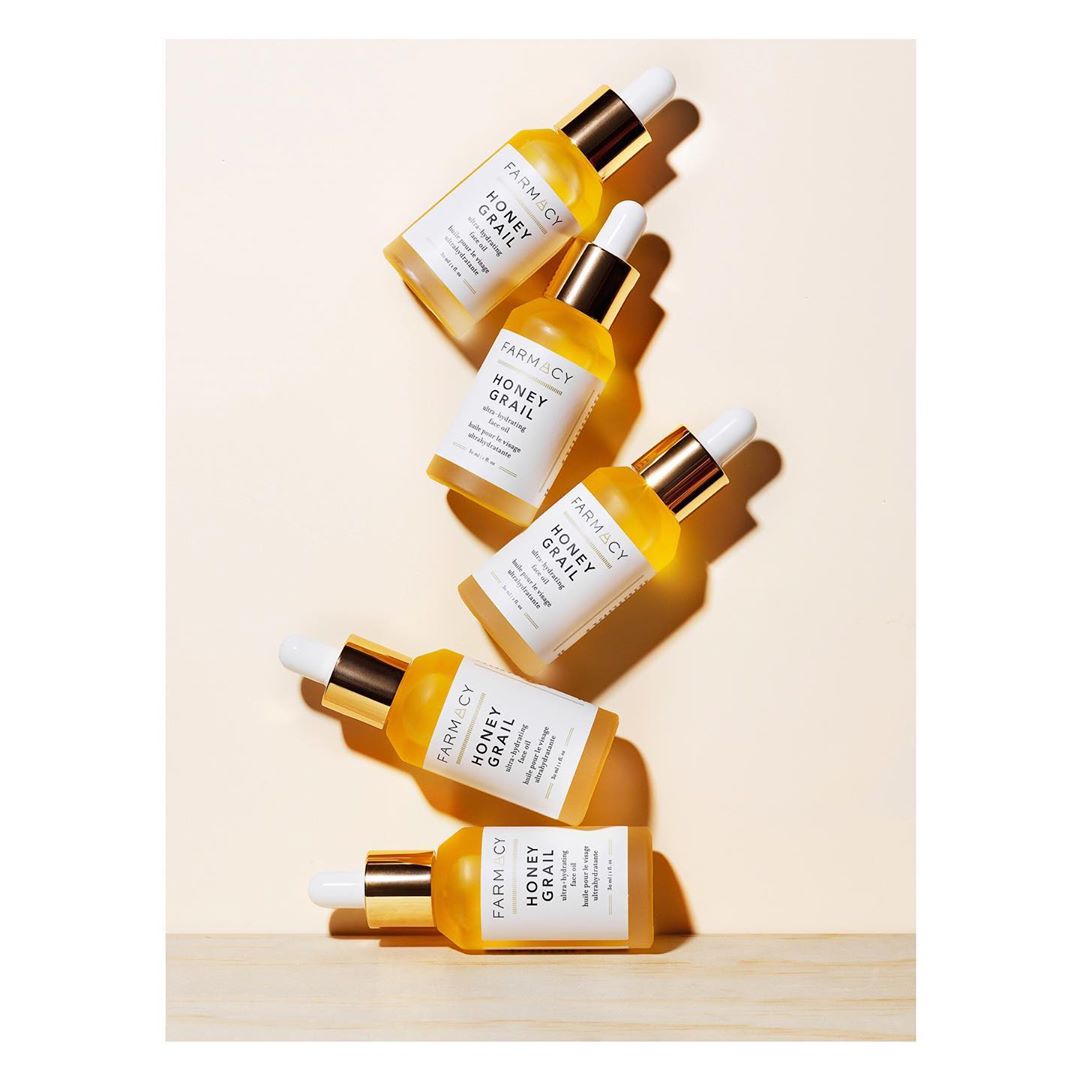 Multiple bottles of Farmacy Honey Grail facial oil with white dropper caps sit on a light-yellow background with a wooden surface.