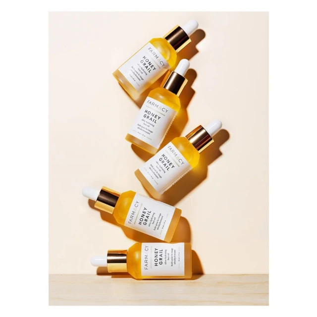 Multiple bottles of Farmacy Honey Grail facial oil with white dropper caps sit on a light-yellow background with a wooden surface.