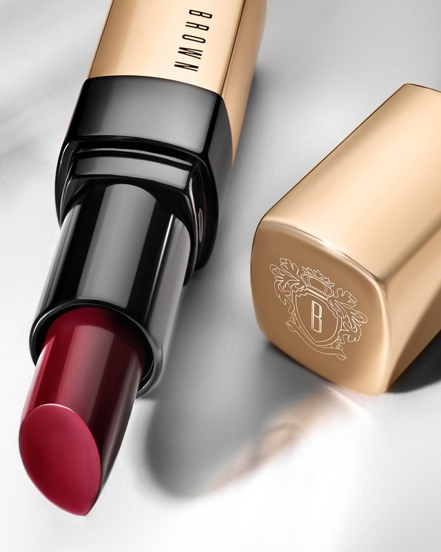 Close-up studio shot of deep-red Bobbi Brown lipstick in a gold and black tube with the lid off, sitting against a grey background.