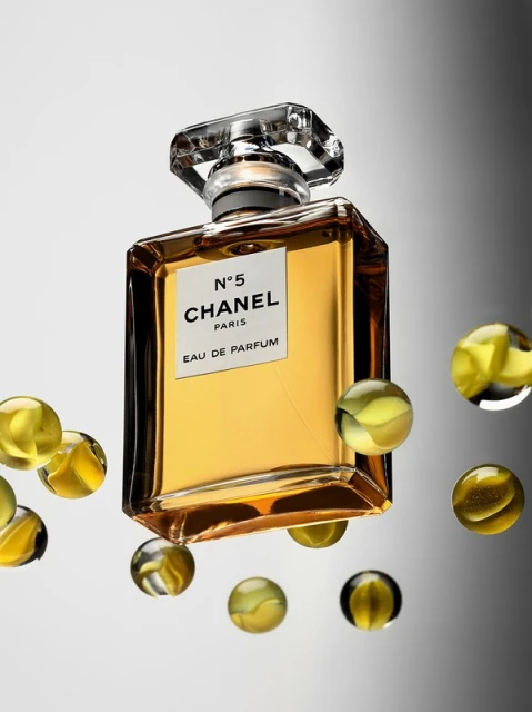 Elegant square perfume bottle with gold liquid and faceted crystal cap, surrounded by translucent yellow-green spheres on gray background.