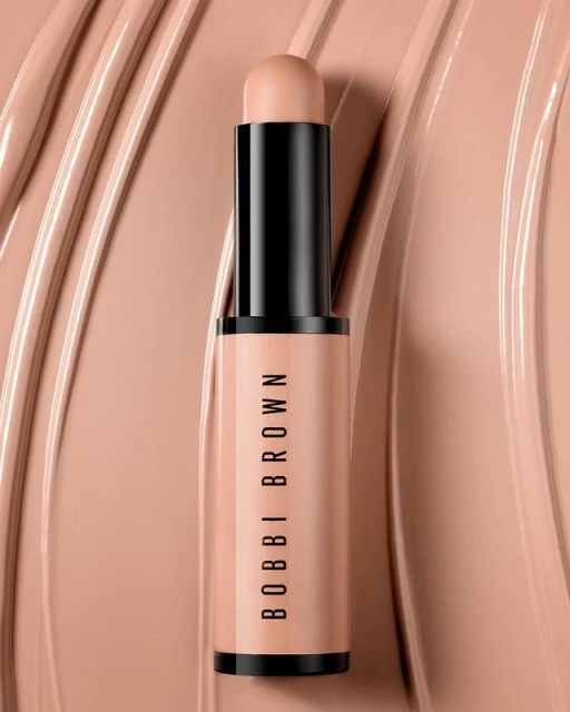 Close-up studio shot of a Bobbi Brown concealer stick. The product, with black accents, sits against a background of matching concealer swirls.