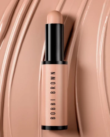 Close-up studio shot of a Bobbi Brown concealer stick. The product, with black accents, sits against a background of matching concealer swirls.