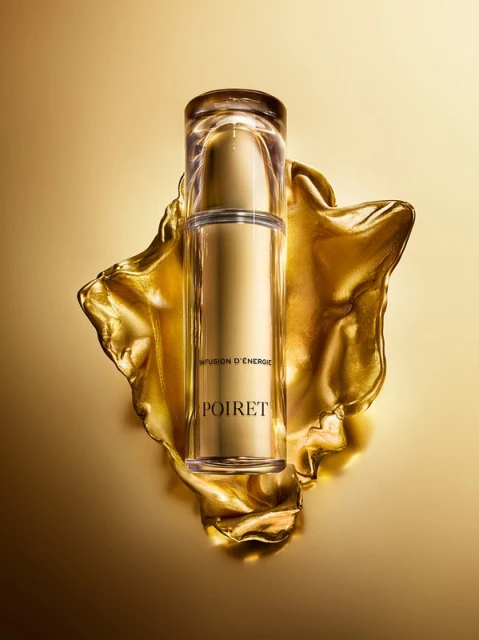 Elevated eye-level studio shot of a gold-toned Poiret serum bottle on a flowing gold fabric with clear cap against a gold background.