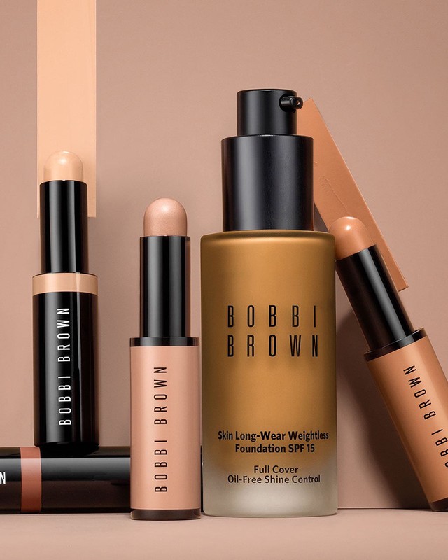 Bobbi Brown makeup products including foundation and concealer sticks in a studio product shot with neutral background and professional lighting.