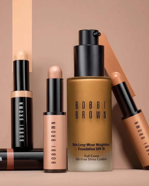 Bobbi Brown makeup products including foundation and concealer sticks in a studio product shot with neutral background and professional lighting.