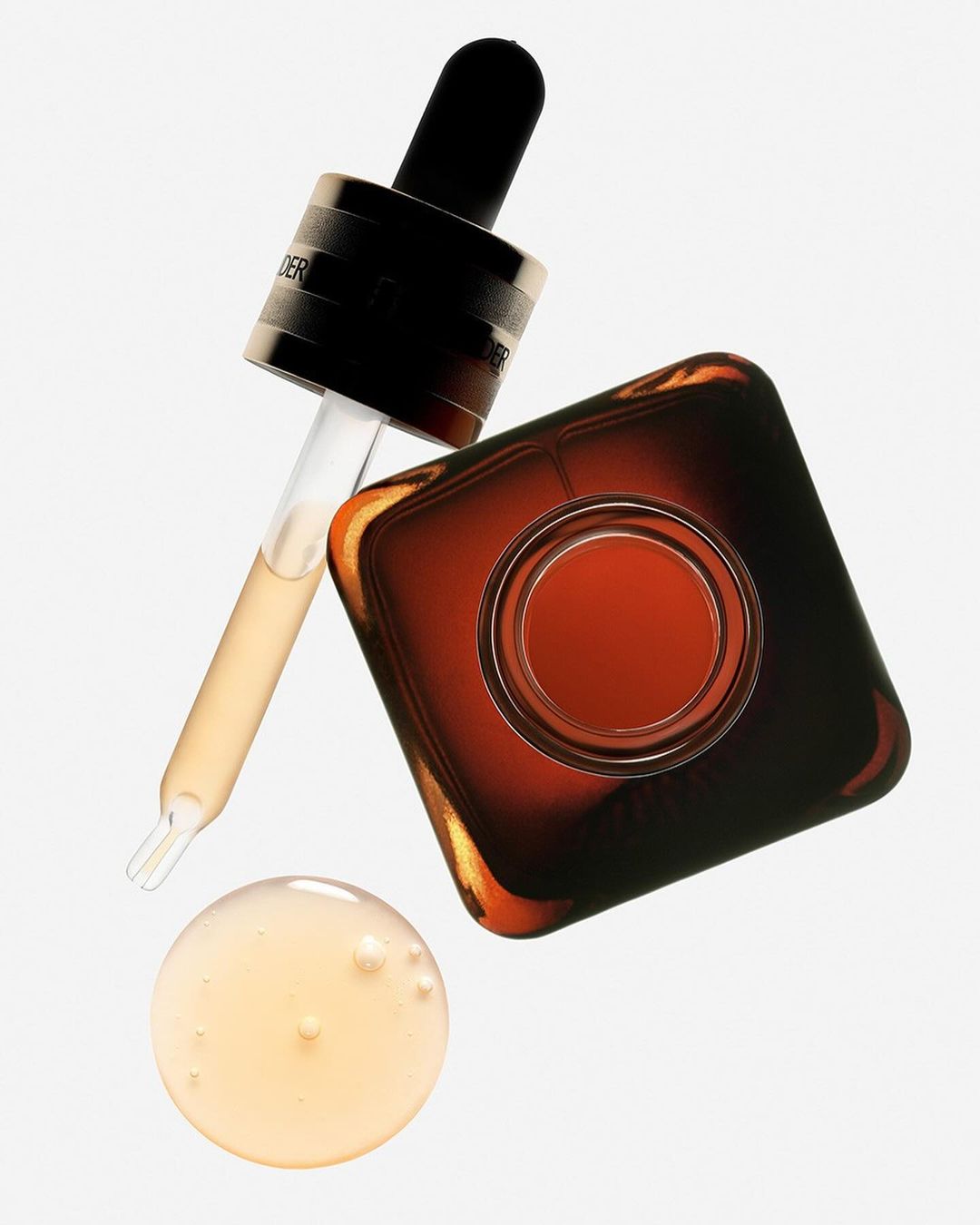 Close-up of amber-colored cosmetic serum in a square bottle with a black dropper and a small puddle of serum.