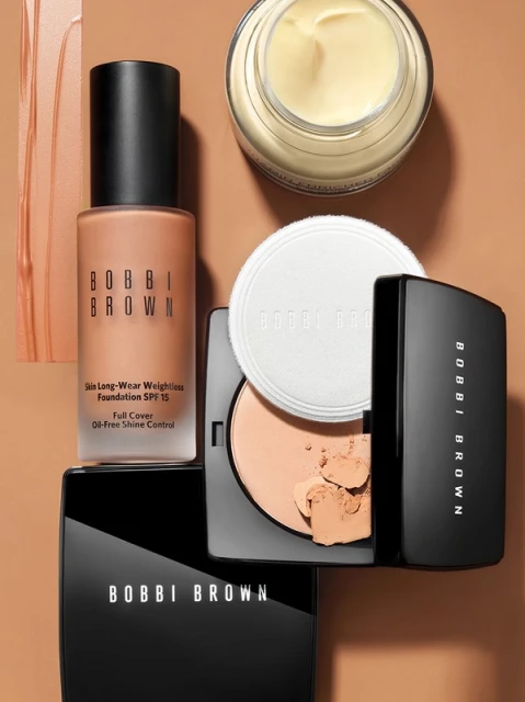Bobbi Brown makeup collection flat lay featuring foundation, powder, and cream products against a warm, neutral backdrop. Studio shot.