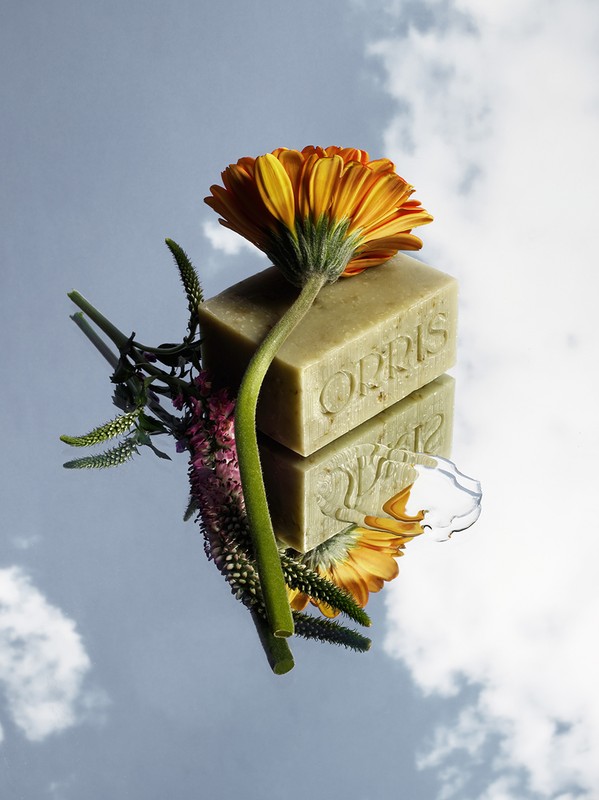 Still life shot of Orris soap and orange gerbera daisy flower lying on a mirror reflecting a blue sky with white clouds.