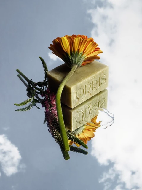 Still life shot of Orris soap and orange gerbera daisy flower lying on a mirror reflecting a blue sky with white clouds.