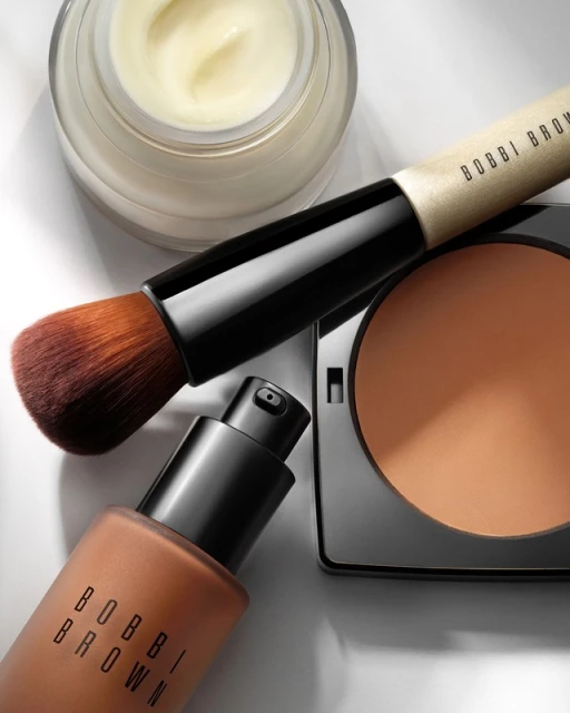 Close-up of Bobbi Brown makeup: foundation bottle, face powder compact, makeup brush, and moisturizer on a clean white surface.
