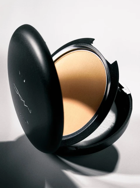 A close-up of a MAC Studio Fix powder foundation compact in a black case with golden powder and a white applicator, on a white background.