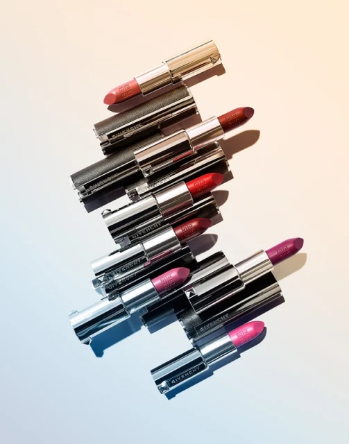 Stack of Givenchy lipsticks in silver tubes with various pink and red shades, arranged on a gradient background, lit for product photography.