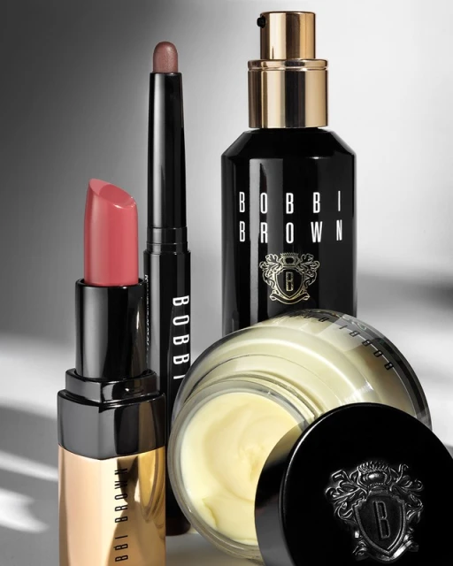 Bobbi Brown makeup products including lipstick, eyeshadow stick, face cream, and serum arranged on a gradient gray background.