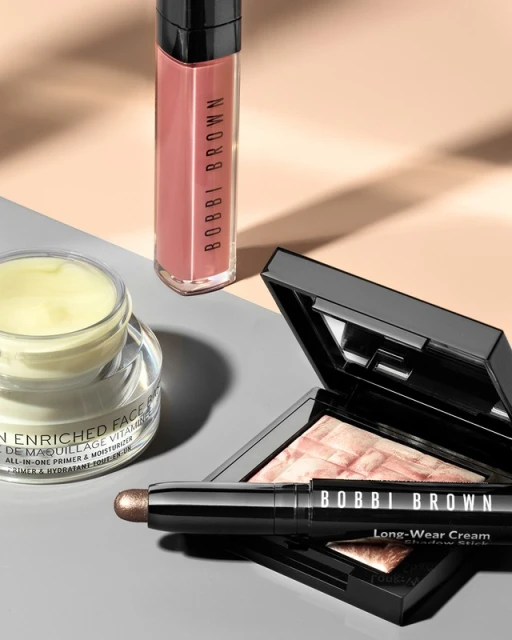 Close-up of Bobbi Brown makeup products including face cream, lip gloss, eye shadow stick and face powder on a neutral surface.