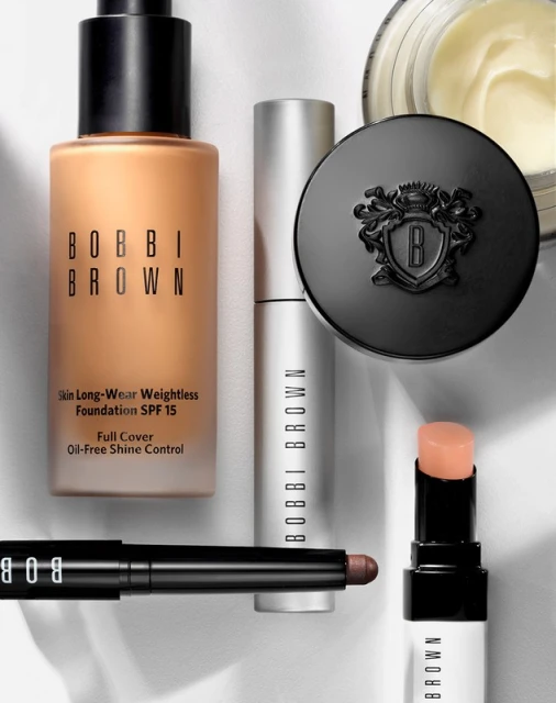 Bobbi Brown makeup products, including foundation, concealer, lip balm, and face powder, are arranged on a white surface.
