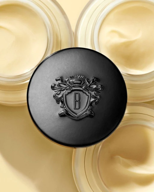 Close-up of three jars of cream, the central jar's lid is emblazoned with a 'B' logo, set against a complementary light-yellow background.