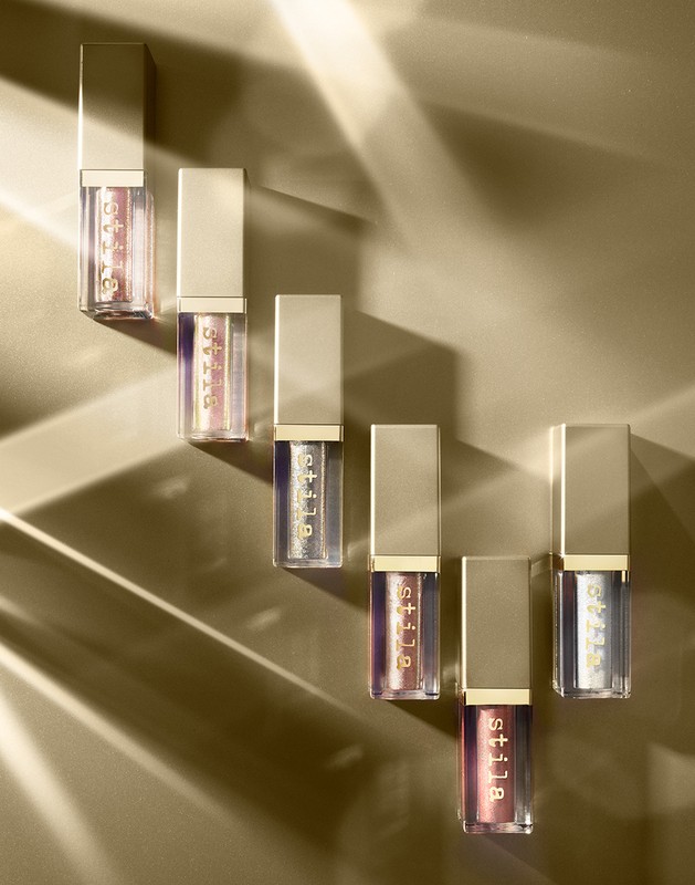 Six bottles of Stila liquid eyeshadow are arranged on a gold background, with light creating a textured shadow across the scene.