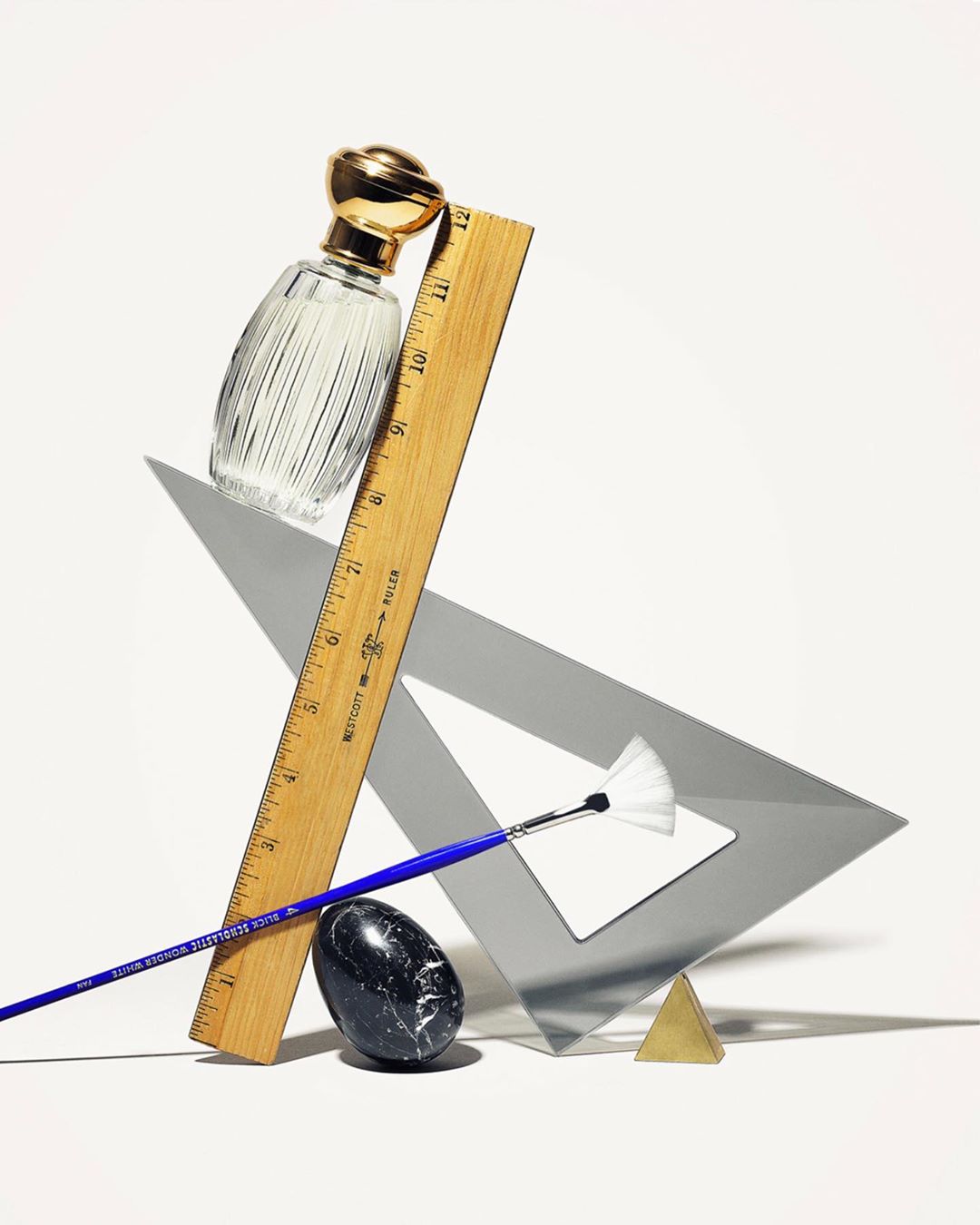 Still life photography featuring a perfume bottle, wooden ruler, paint brush, triangular drafting tool, and a dark stone on a white background.