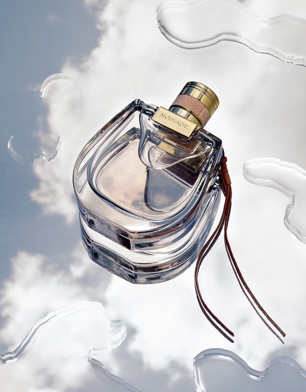 A glass perfume bottle of 'Nomade' is captured floating on a reflective surface with water droplets, against a soft sky background.