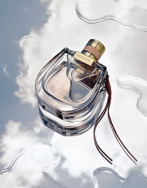 A glass perfume bottle of 'Nomade' is captured floating on a reflective surface with water droplets, against a soft sky background.