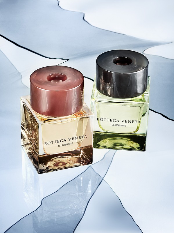 Two Bottega Veneta Illusione fragrance bottles, one with a rose gold cap, the other with a dark grey cap, against a patterned backdrop.