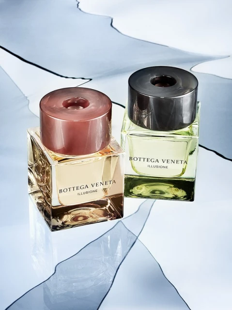 Two Bottega Veneta Illusione fragrance bottles, one with a rose gold cap, the other with a dark grey cap, against a patterned backdrop.