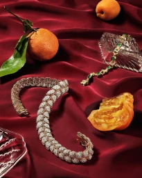 Diamond bracelets, crystal glassware, and fresh citrus fruits arranged on deep red draped silk fabric.