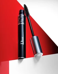 Black Dior mascara tube standing upright with its brush wand propped open against a geometric red and white background