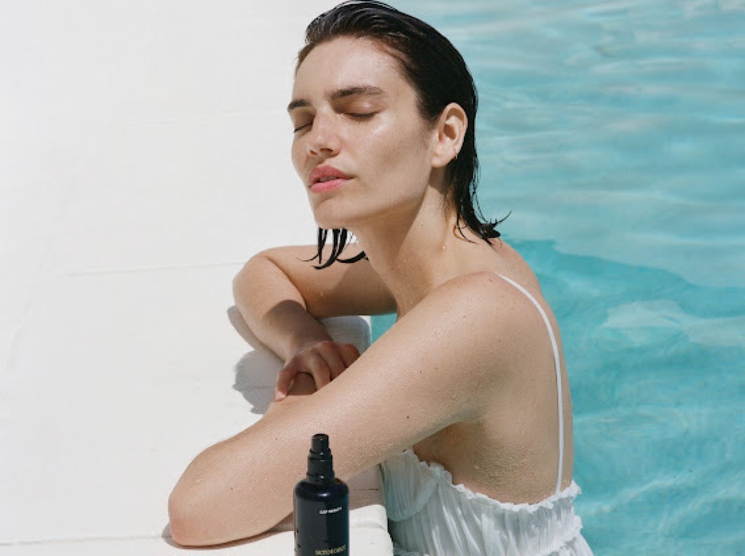 Adult woman relaxing by swimming pool with eyes closed, wearing a white dress. A bottle of product is on the pool ledge.