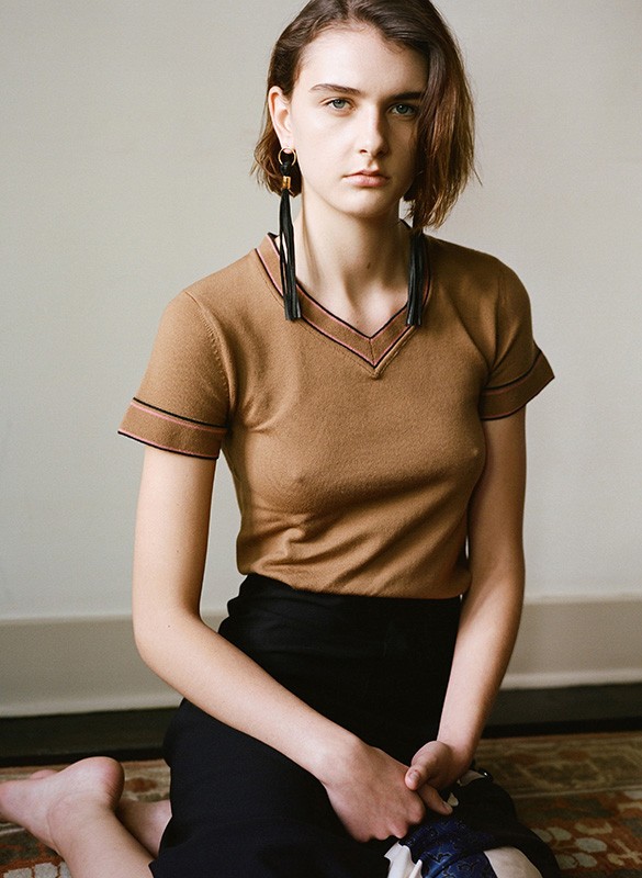 A model kneeling indoors wearing a tan short-sleeve shirt with a black pencil skirt and long, black earrings in a portrait-style photograph.