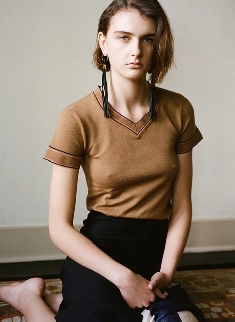 A model kneeling indoors wearing a tan short-sleeve shirt with a black pencil skirt and long, black earrings in a portrait-style photograph.