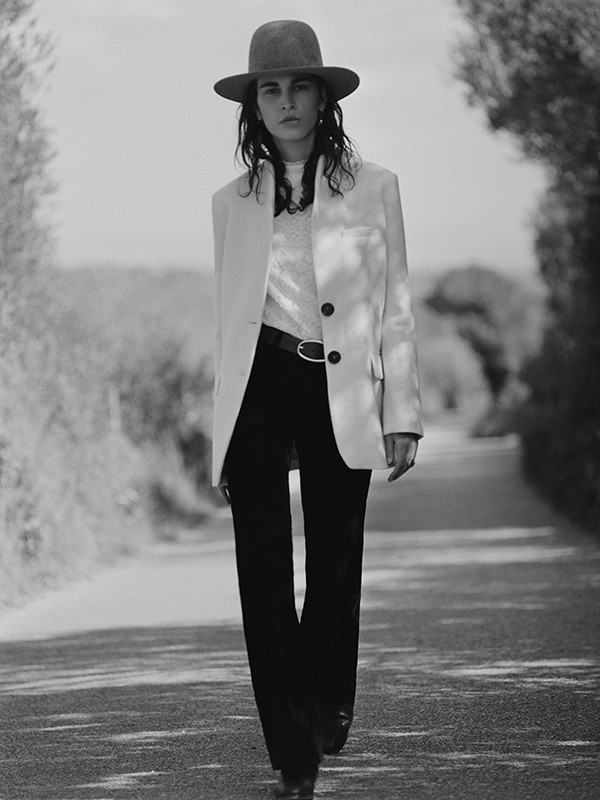 Model in wide-brimmed hat, light blazer, and dark trousers walking along an outdoor tree-lined road in a monochrome portrait.