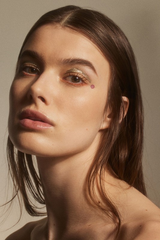 Close-up beauty shot of a young adult female model with light skin, brown hair, and sparkly gold eye makeup in a studio setting.