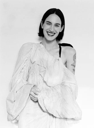 Monochrome portrait of a smiling woman with dark bobbed hair wearing a textured off-shoulder dress against a white background