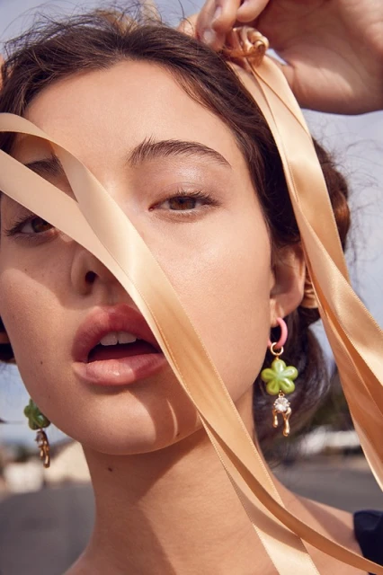 Close-up of a young woman with fair skin and brown hair tying a beige ribbon in her hair, wearing flower earrings with gem pendants.
