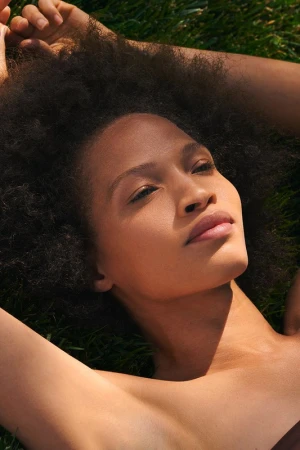 Close-up of an adult woman laying in the grass with her arms above her head, natural light, and curly hair in a portrait orientation.